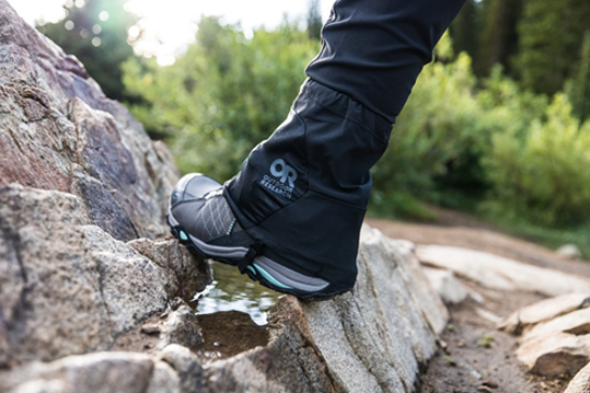 Person wearing sneakers, black hiking pants and a outdoor research gaiter on their foot steps onto a rock along a wilderness trail.