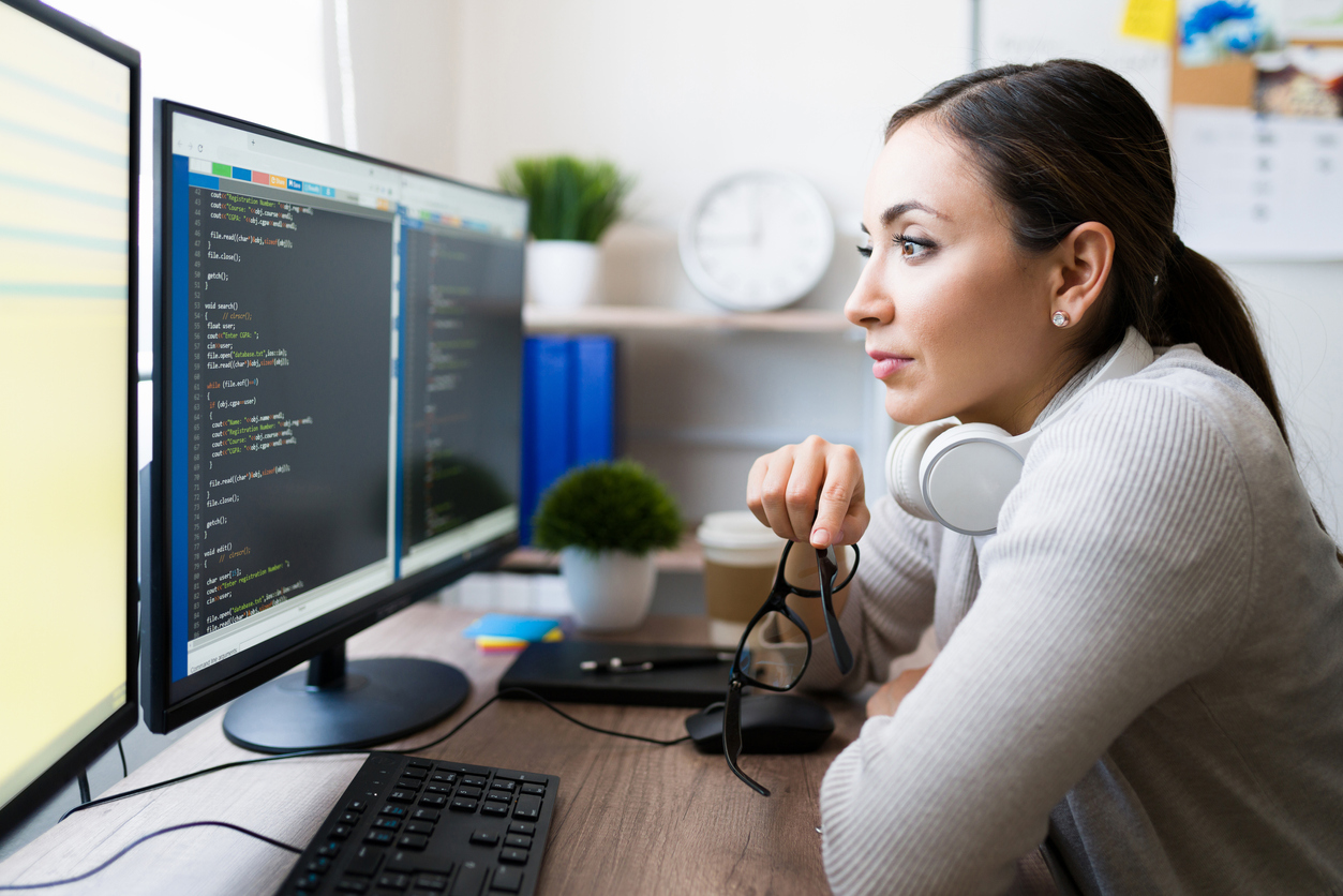 Mujer atractiva de unos 20 años sentada frente a su escritorio y quitándose las gafas mientras lee el código del software