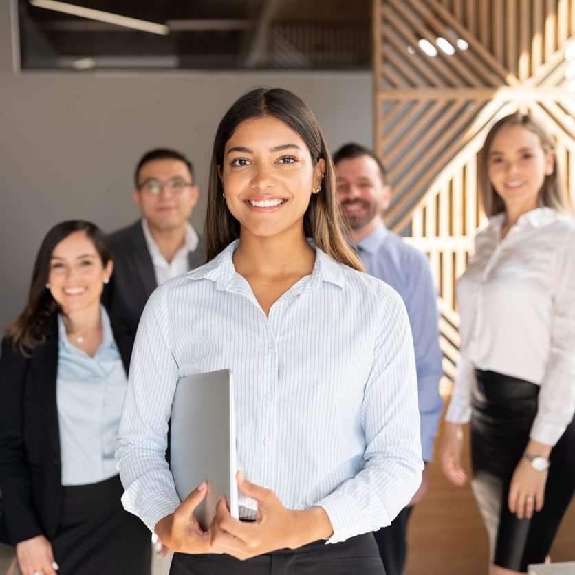 Confident hispanic businesswoman standing in office with colleagues in background